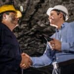 A miner shakes hands with a businessman or banker inside an underground mine setting.