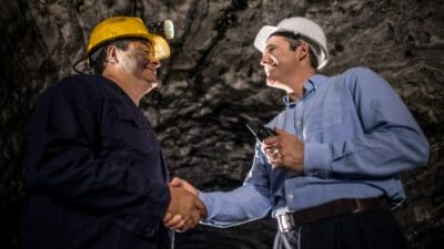 A miner shakes hands with a businessman or banker inside an underground mine setting.