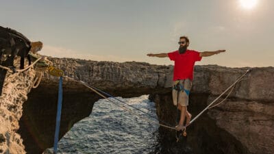 A man balances on a tightrope across rocks above the sea at sunset.