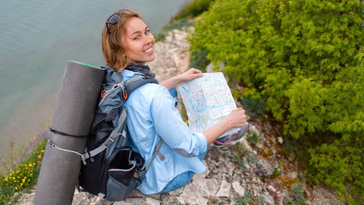 A smiling woman with backpack and a map sits on a rocky cliff about to embark on a new investing journey.