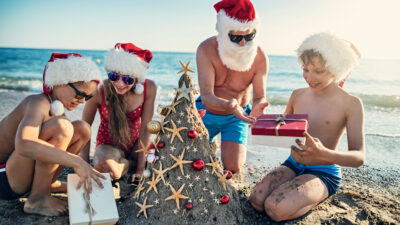 A family of four wearing Santa hats open presents on the beach next to a Christmas tree.