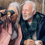 A happy elderly couple enjoy a cuppa outdoors as the woman looks through binoculars.