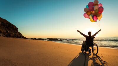A joyful woman in a wheelchair on a beach holds a bunch of colourful balloons and spreads her arms wide towards the sunset.