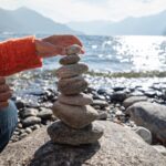 A woman stacks smooth round stones into a pile by a lake.