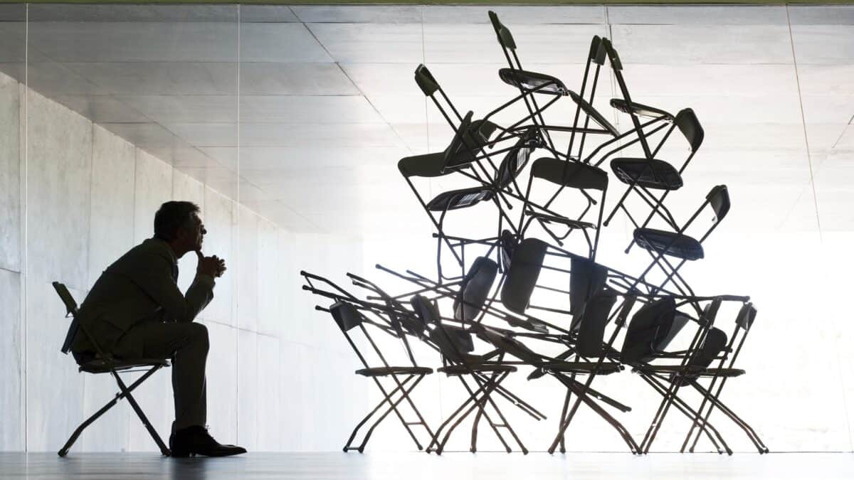 A businessman sits on a chair looking at a pile of chairs stacked up to the ceiling of a white empty room.