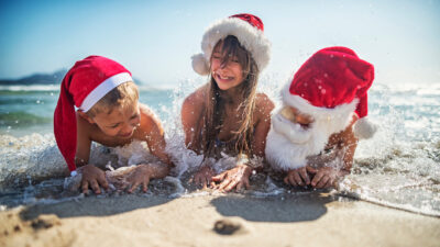 Three young people lie in the surf on a beach wearing santa hats.