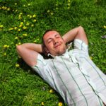 A happy, smiling man stretches out among yellow daisies in the green grass, dreaming of success.