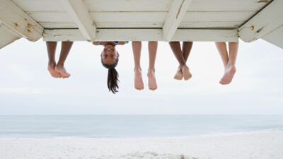 Five friends sit on the end of a jetty, four with feet dangling and the fifth upside down smiling at the camera.