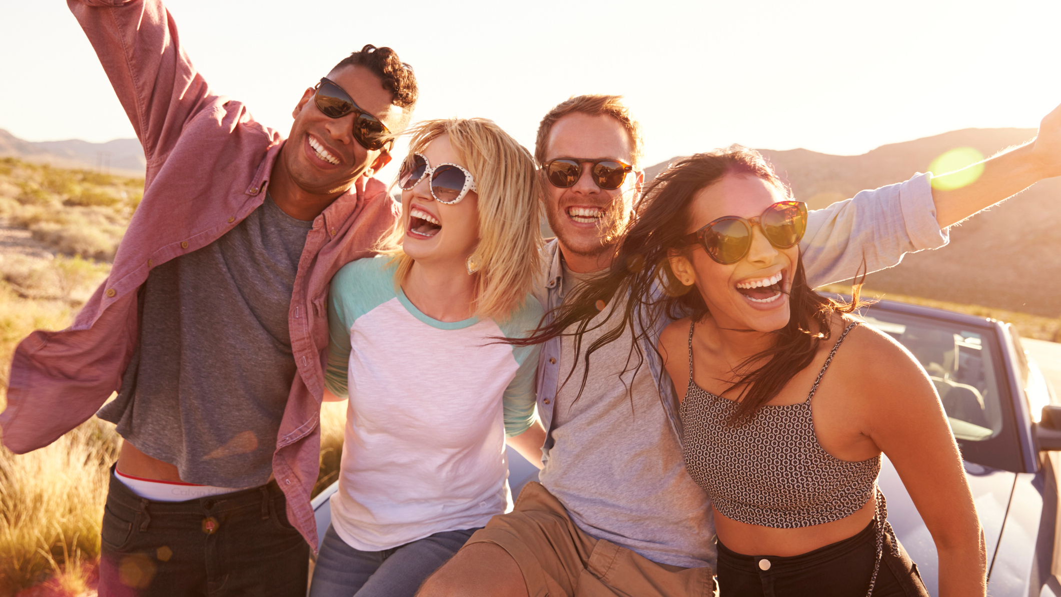 Four young friends on a road trip smile and laugh as they sit on roof of their car.