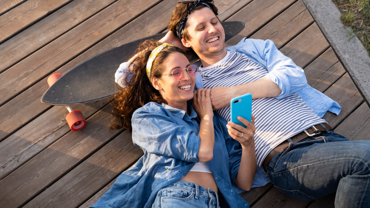 A happy young couple lie on a wooden deck using a skateboard for a pillow.
