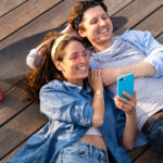 A happy young couple lie on a wooden deck using a skateboard for a pillow.