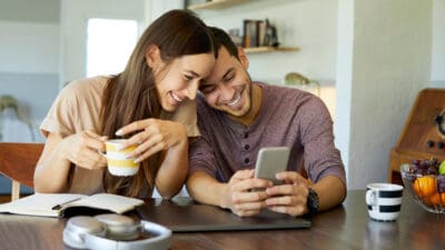 Cheerful boyfriend showing mobile phone to girlfriend in dining room. They are spending leisure time together at home and planning their financial future.