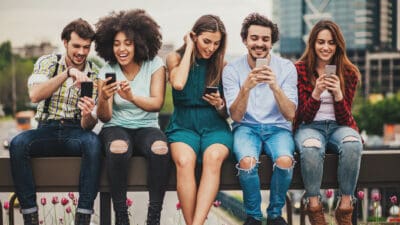 Five young people sit in a row having fun and interacting with their mobile phones.