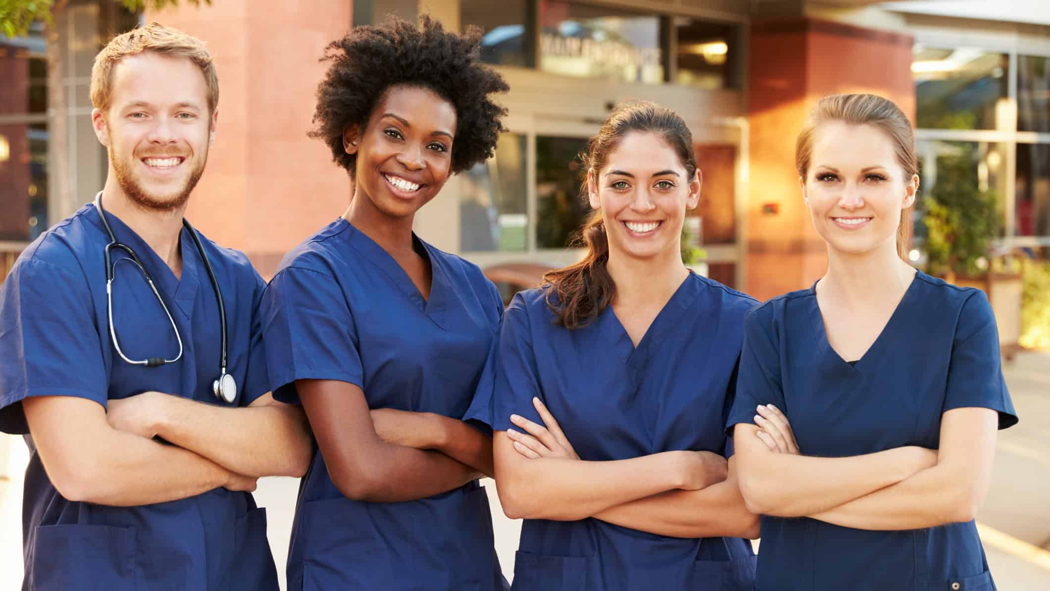 Four smiling young medics with arms crossed stand outside a hospital.