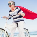 A mature-aged woman wearing goggles and a red cape, rides her bike along the beach looking victorious.