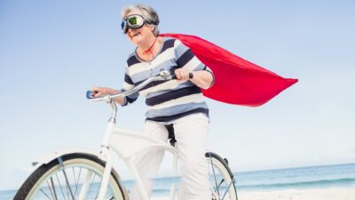 A mature-aged woman wearing goggles and a red cape, rides her bike along the beach looking victorious.
