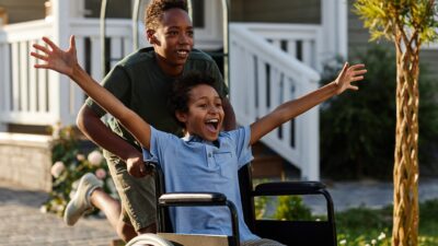 A happy young boy in a wheelchair holds his arms outstretched as another boy pushed him.