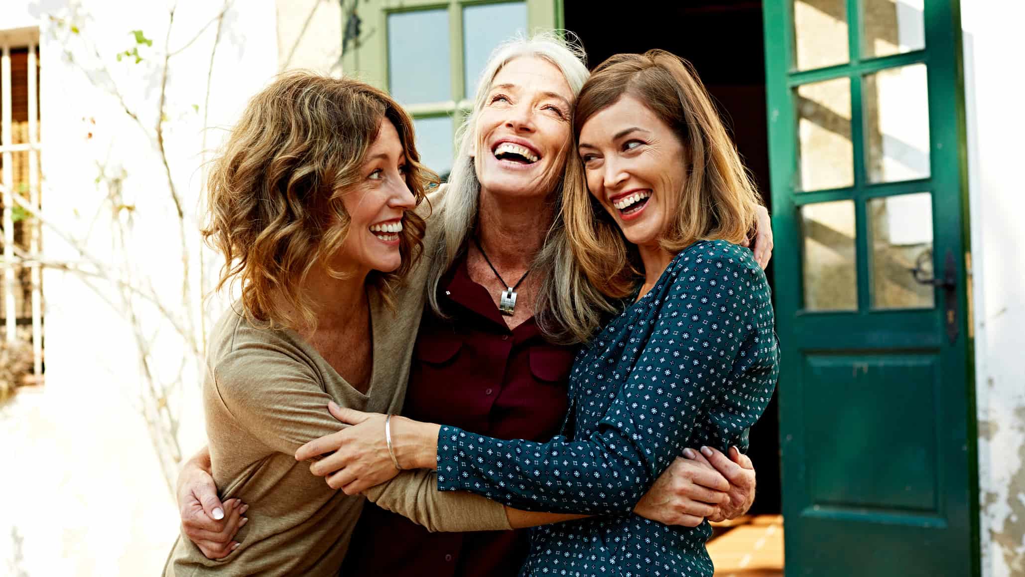 A mother and her two adult daughters embrace outdoors.