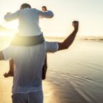 A young boy sits on his father's shoulders as they flex their muscles at sunrise on a beach