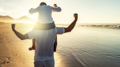 A young boy sits on his father's shoulders as they flex their muscles at sunrise on a beach