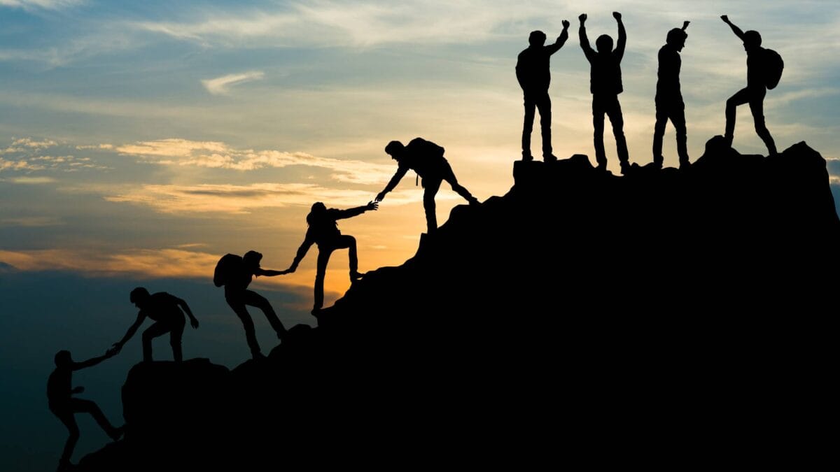 Silhouettes of nine people climbing a steep mountain to the top at sunset, and helping each other along the way.