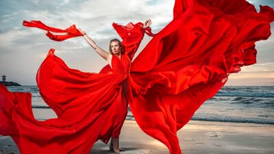 A woman wearing a flowing red dress, poses dramatically on a beach with the sea in the background.