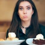 A bemused woman tries to choose between two slices of cake she holds on two plates.