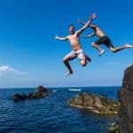 Two young risk-taking men pose for the camera as they jump off a cliff into the sea.