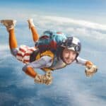 A colourfully dressed young skydiver wearing heavy gold gloves smiles and gives a thumbs up as he falls through the sky.