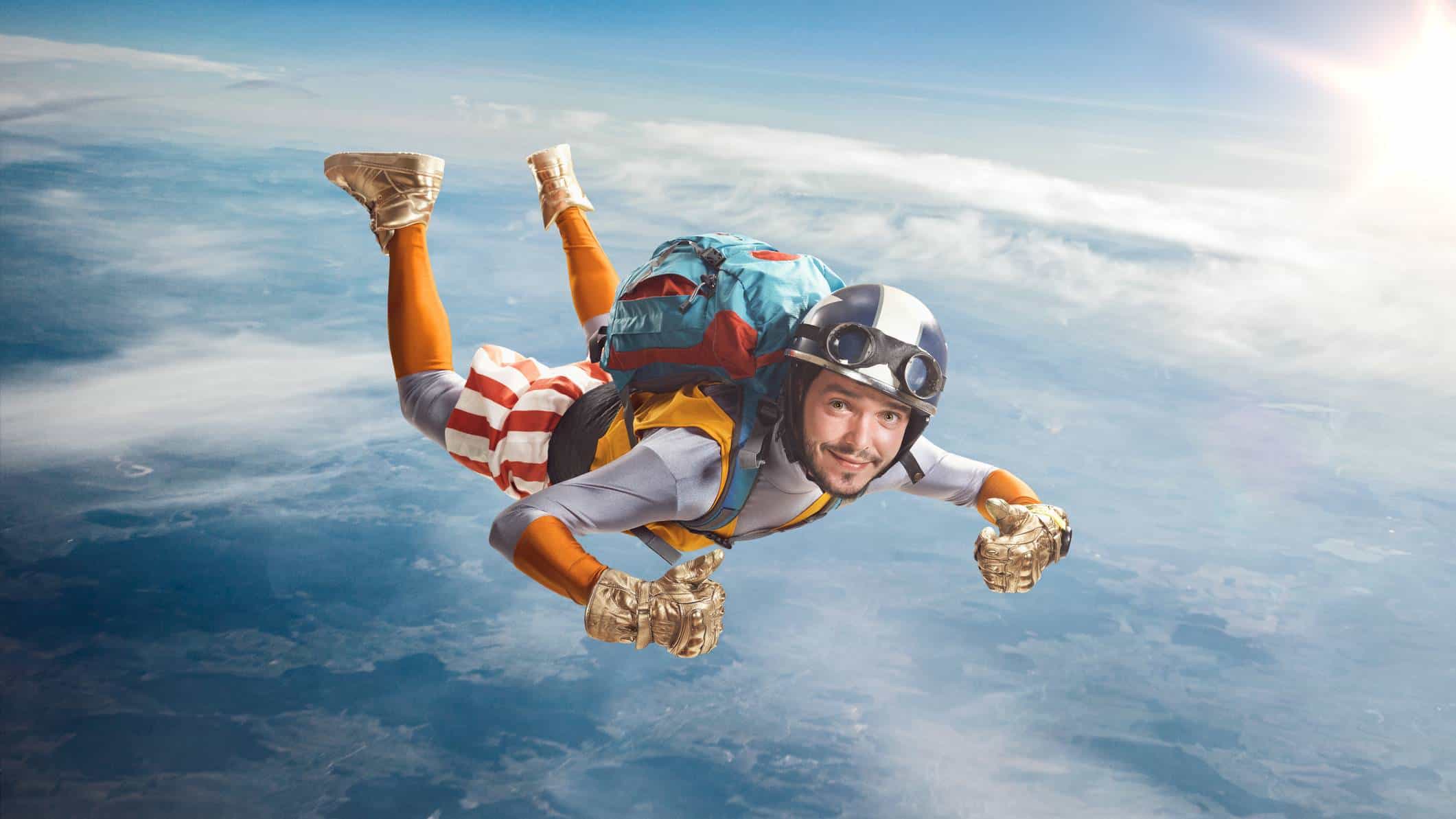 A colourfully dressed young skydiver wearing heavy gold gloves smiles and gives a thumbs up as he falls through the sky.