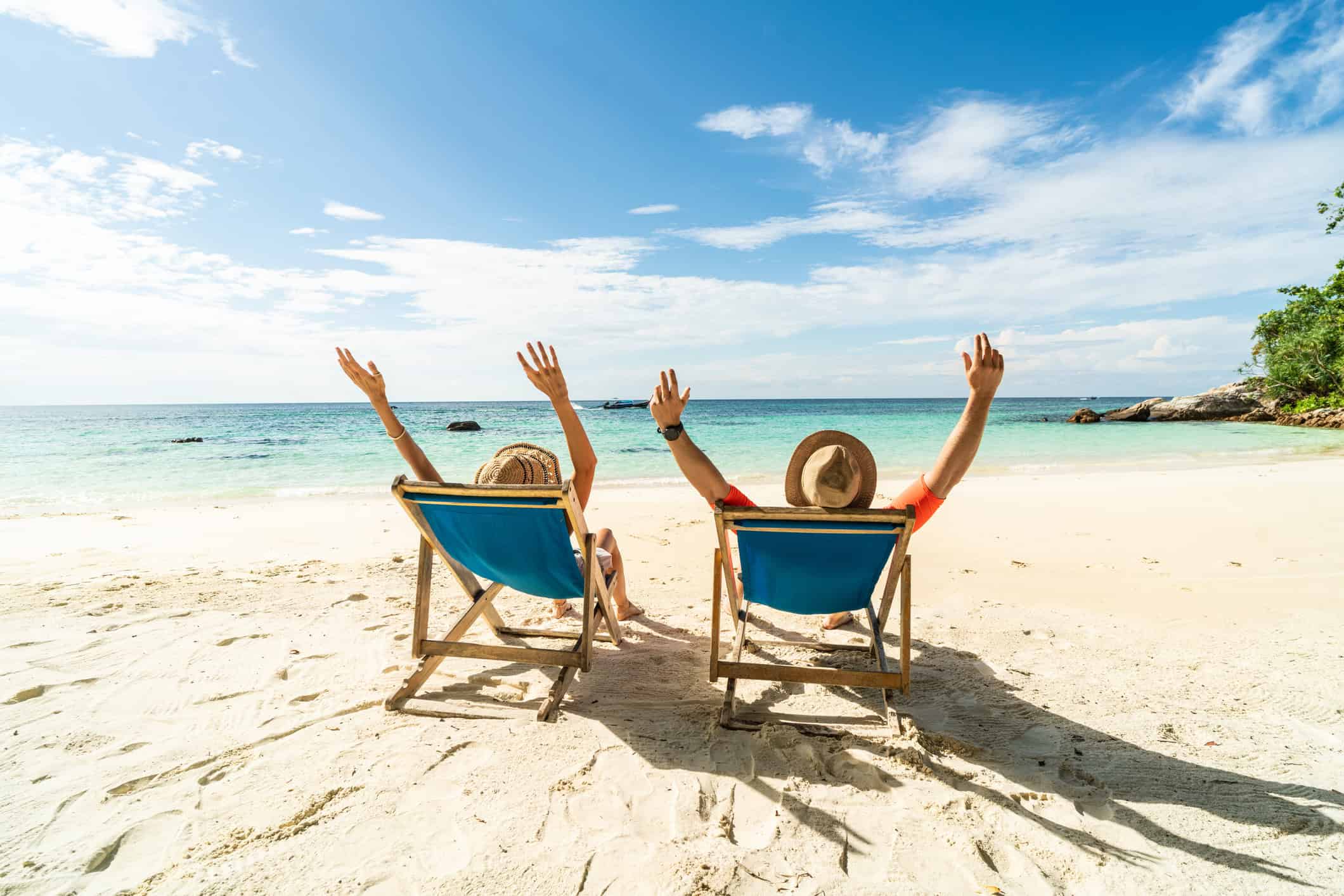 Two people lazing in deck chairs on a beautiful sandy beach throw their hands up in the air.