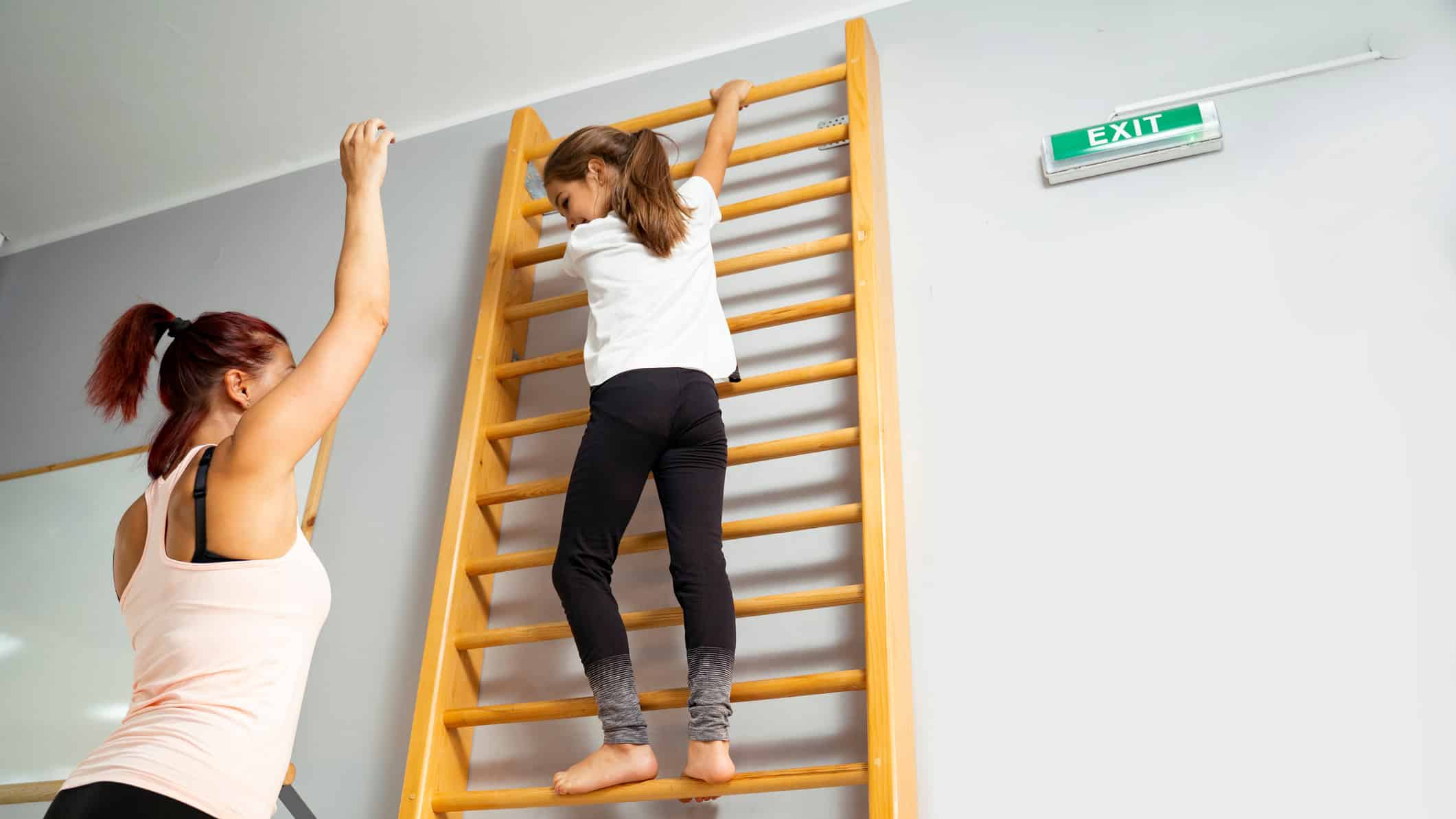 Sport trainer talking to little girl who is climbing wooden ladder in gym.