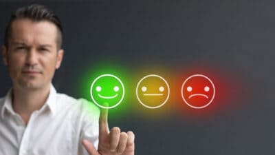 Man pressing smiley face emoji on digital touch screen next a neutral faced and sad faced emoji.