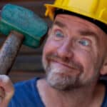 A man holds a green hammer while wearing a yellow hardhat.