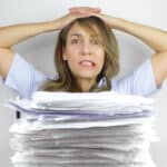A business woman looks frustrated and angry at a huge stack of paperwork on her desk.