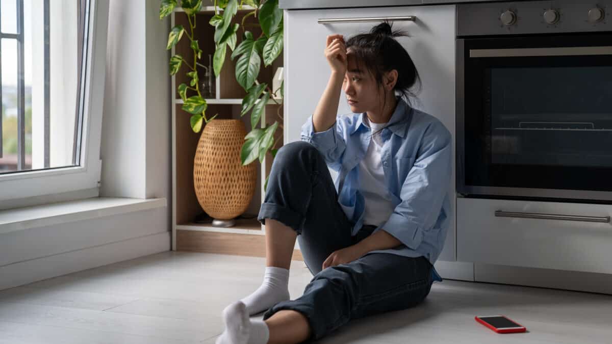 Devastated woman sits near smartphone on home kitchen floor troubled with loneliness.