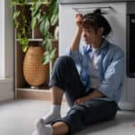 Devastated woman sits near smartphone on home kitchen floor troubled with loneliness.