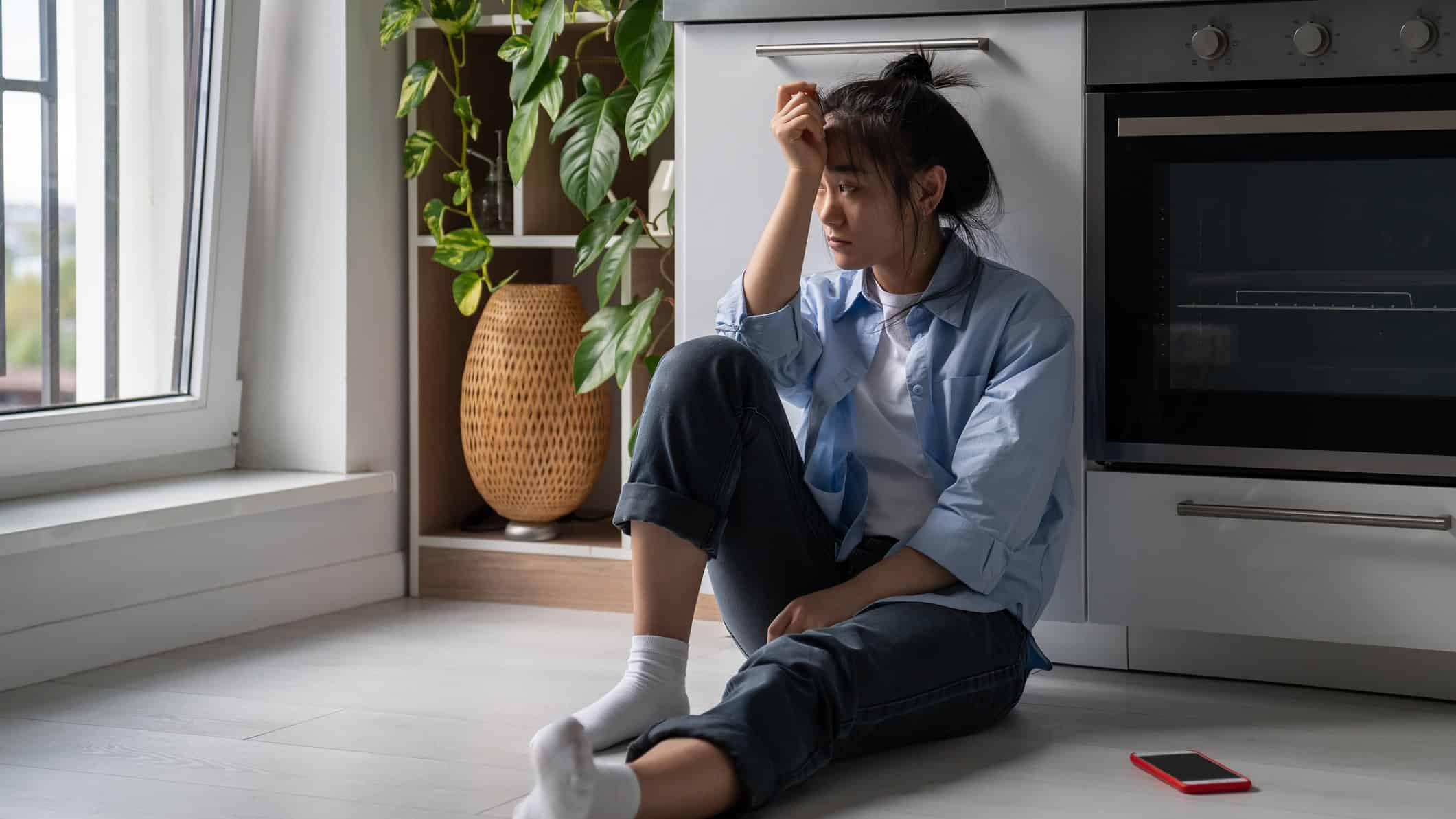 Devastated woman sits near smartphone on home kitchen floor troubled with loneliness.
