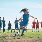 A player kicks a soccer ball to score a goal while players from both teams watch on.