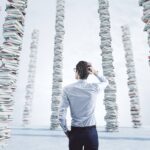 A geeky business man scratches his head as he looks at many stacks of books piled up on the floor.