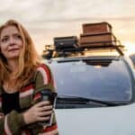 A woman smiles as she stands next to a car loaded with a stack of suitcases on the roof.