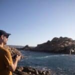 A man wearing a cap flies his drone at the beach.