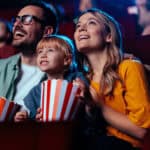 A young joyful couple is watching a movie with their daughter in the cinema.