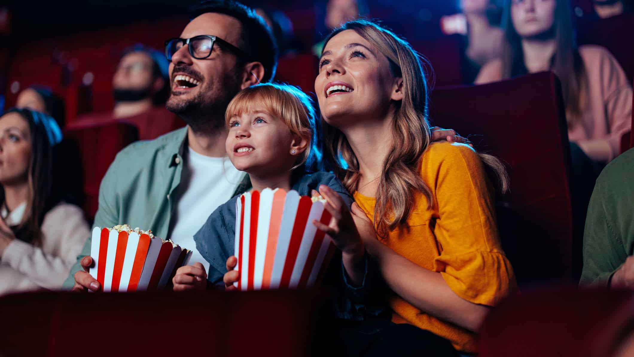A young joyful couple is watching a movie with their daughter in the cinema.