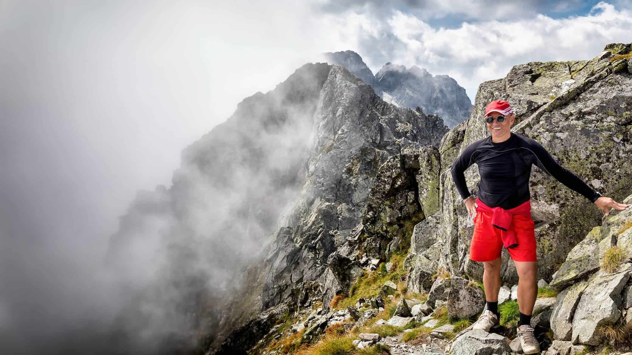 Man smiling on top of rocks with mountains in the background.