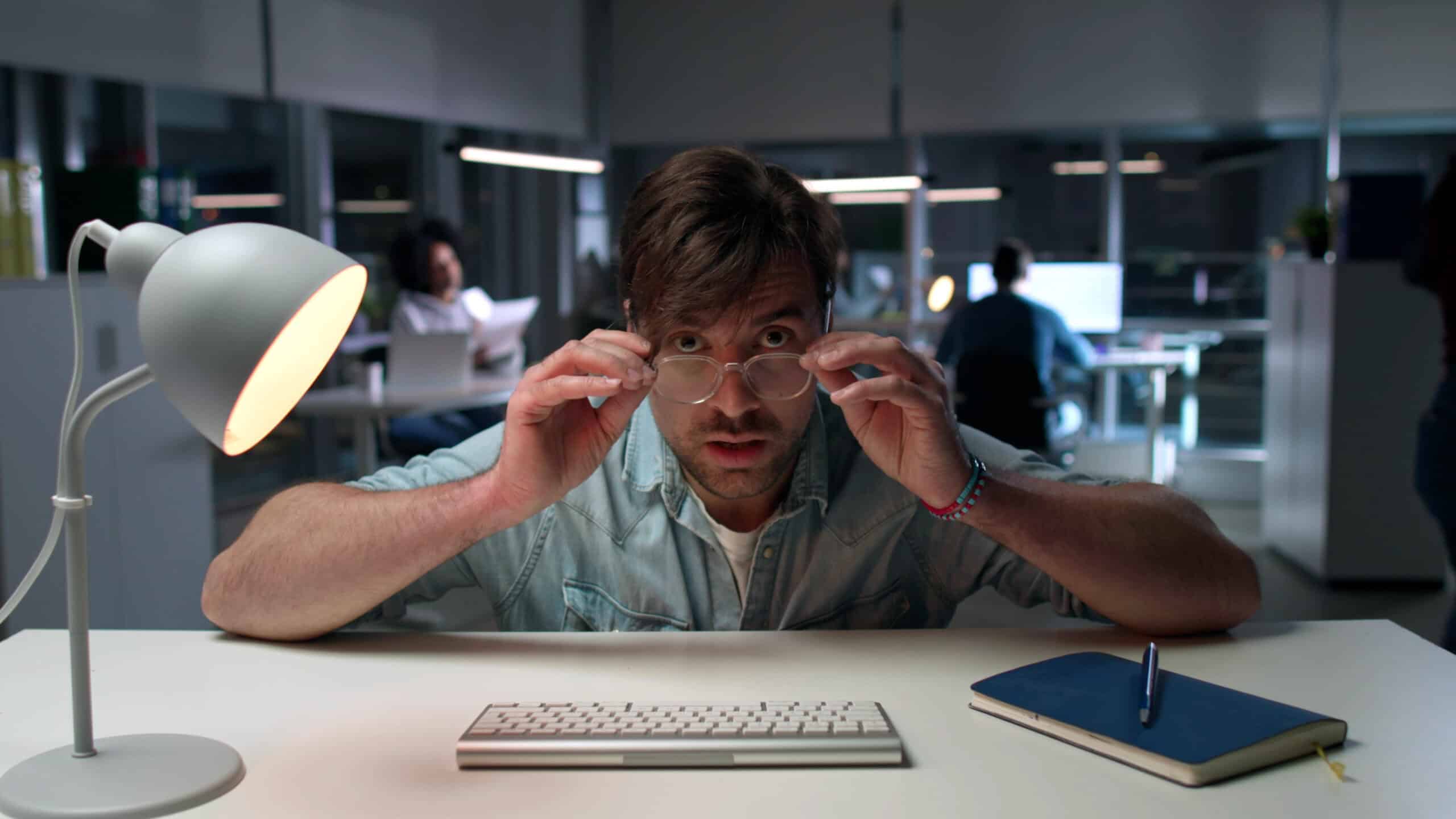 Shocked office worker staring at computer screen with colleagues working in the background.