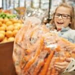 A happy youngster holds a giant bag of carrots at a supermarket fruit and vegie section, indicating savings made by buying in bulk.