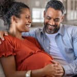 Happy father hands on expecting mother's baby bump while embracing her on couch.