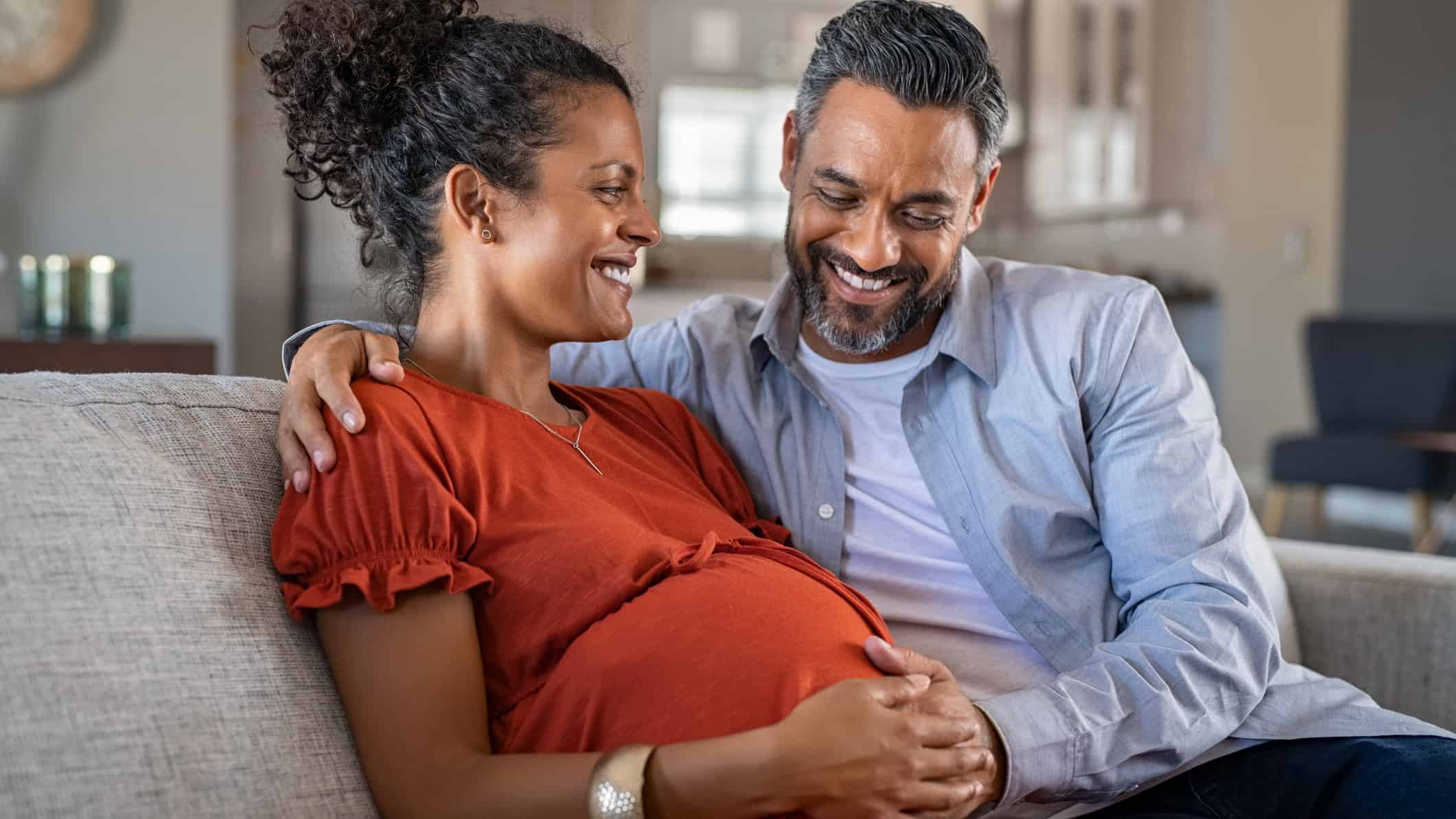Happy father hands on expecting mother's baby bump while embracing her on couch.