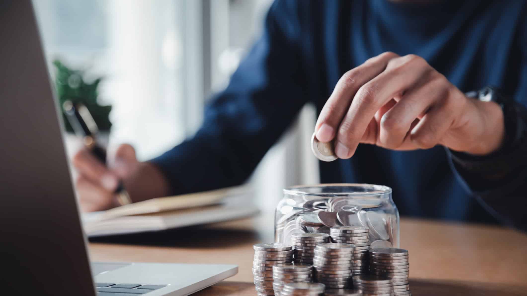 Man putting in a coin in a coin jar with piles of coins next to it.